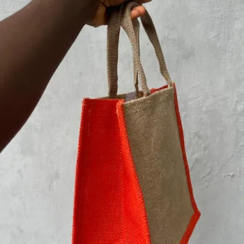 Handmade jute tote bag with orange side panels and natural beige body, held by hand against a grey wall