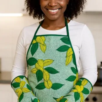 Smiling woman in a kitchen wearing a green and yellow floral apron with matching oven mitts, cutting tomatoes.