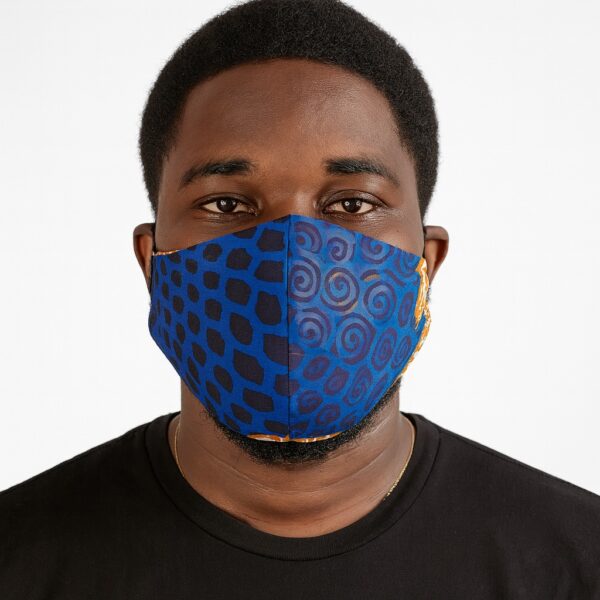 Close-up of a man wearing a blue Ankara face mask with mixed patterns of black animal spots and blue spiral motifs. Clean white background, front-facing view.
