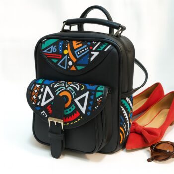 Black leather backpack with colorful Ankara patterns beside red flats and sunglasses in a bright studio setting.