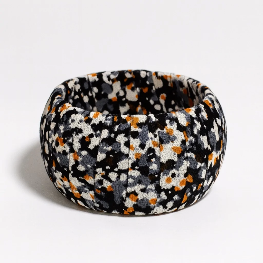 Ankara print bangle with abstract black, white, orange, and gray speckles on fabric, shot on a white background.