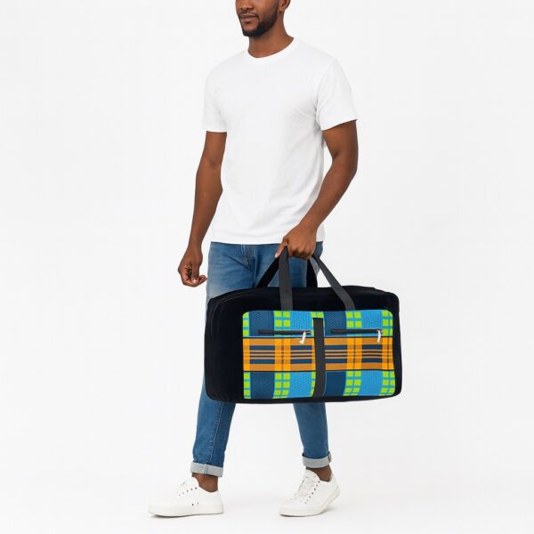 A male model in casual white T-shirt and jeans carrying a large collapsible African print duffel bag by the handles. The bag features a bold checkered Ankara pattern with zippered pockets and sturdy black canvas structure.