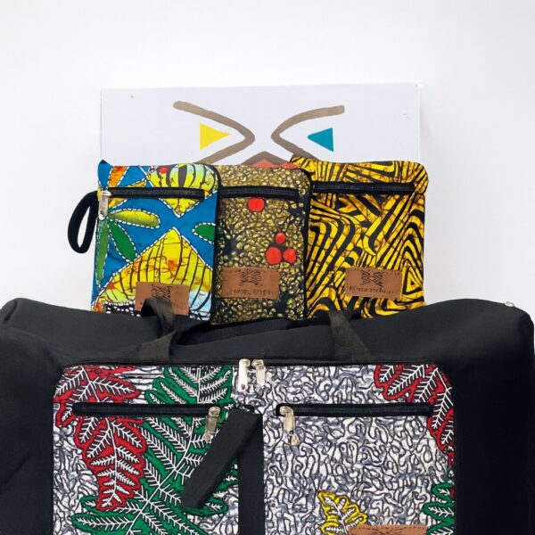 Collection of collapsible African print duffel bags and matching pouches in various bold patterns, including blue, red, and yellow motifs. Displayed upright against a white wall, emphasizing vibrant color combinations and modern cultural styling.