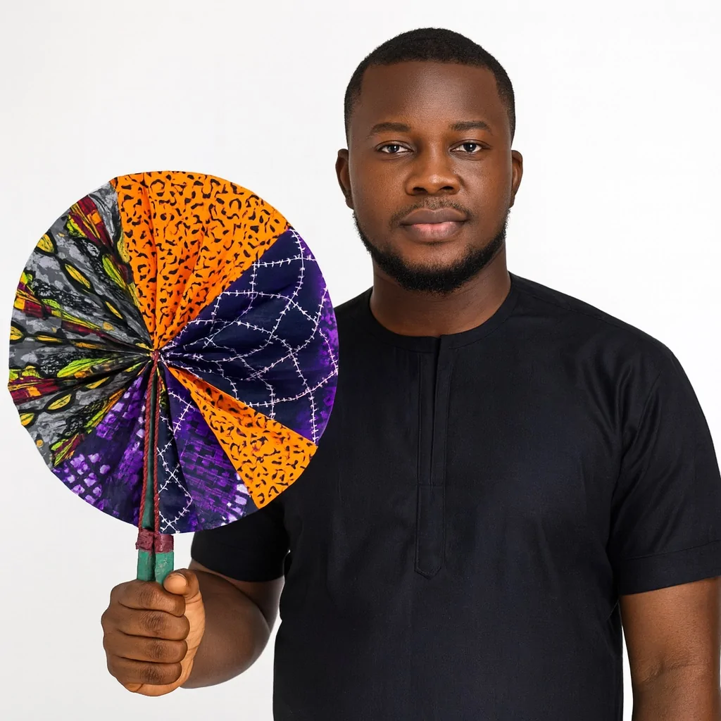 Confident African man in a black traditional outfit holding a vibrant hand fan with mixed Ankara prints in purple, orange, yellow, and grey.