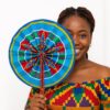 Smiling African woman holding a blue African print hand fan that covers half her face. She wears a matching Kente-style off-shoulder top against a white background.