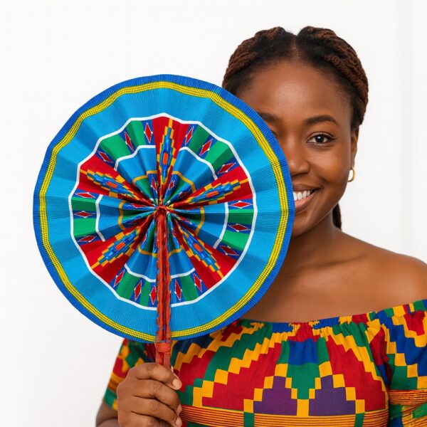 Smiling African woman holding a blue African print hand fan that covers half her face. She wears a matching Kente-style off-shoulder top against a white background.