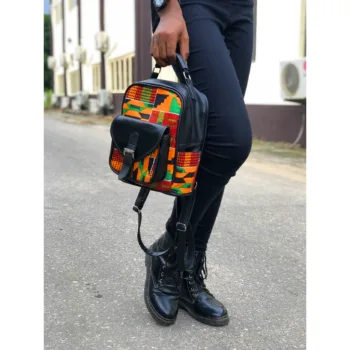 Small African print backpack carried by hand – A stylish urban street-style shot of a small Kente-pattern backpack with black leather accents, held by a person dressed in black pants and lace-up boots while standing outdoors.