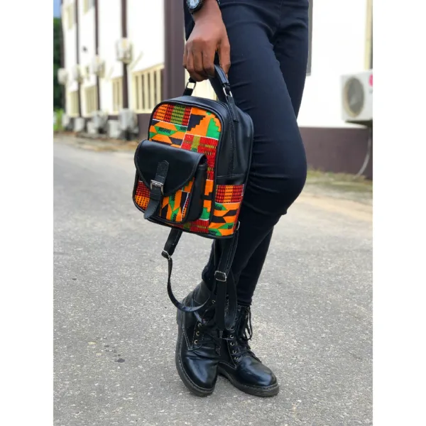 Small African print backpack carried by hand – A stylish urban street-style shot of a small Kente-pattern backpack with black leather accents, held by a person dressed in black pants and lace-up boots while standing outdoors.