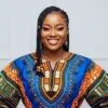 A confident and smiling young Nigerian woman. She is standing against a clean, minimalist background in a well-lit studio. She is wearing a modern, exquisitely tailored dashiki top made from a bold Ankara print with electric blue and sunset orange patterns.