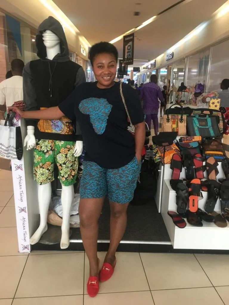 A lady with a black t-shirt carry an African logo, and African print shorts, standing in a shop.