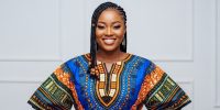 A confident and smiling young Nigerian woman. She is standing against a clean, minimalist background in a well-lit studio. She is wearing a modern, exquisitely tailored dashiki top made from a bold Ankara print with electric blue and sunset orange patterns.