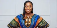 A confident and smiling young Nigerian woman. She is standing against a clean, minimalist background in a well-lit studio. She is wearing a modern, exquisitely tailored dashiki top made from a bold Ankara print with electric blue and sunset orange patterns.